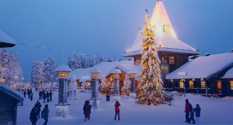 Winter wonderland with people, snow, Christmas tree, lights, and festive wooden buildings.