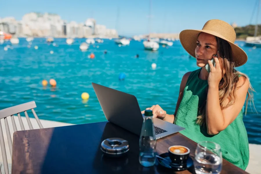 Woman phone-laptop work by ocean café, cityscape background, relaxing modern digital nomad lifestyle.