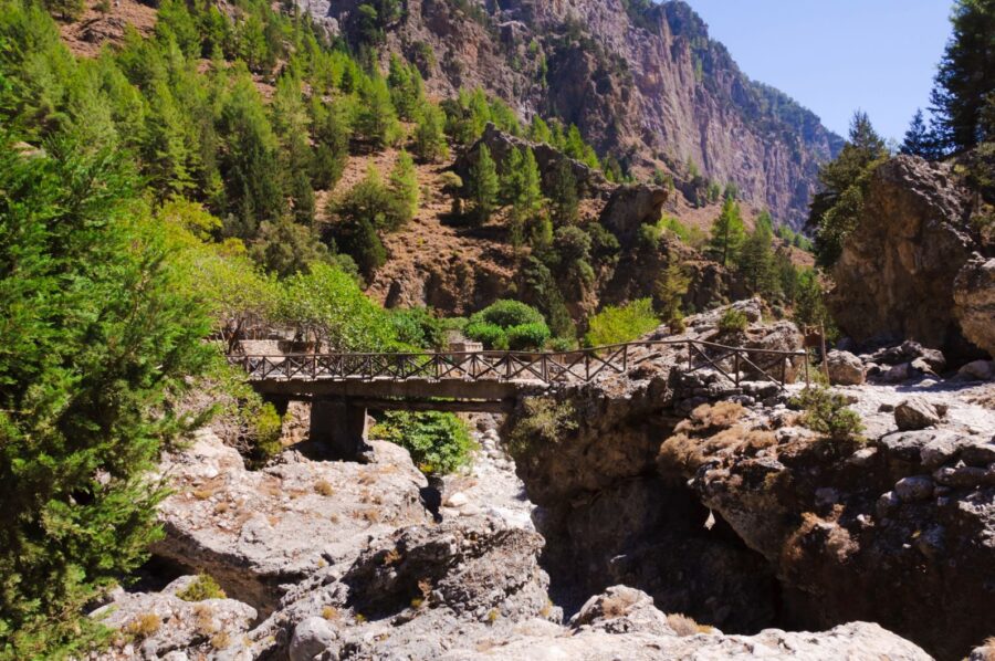 Wooden bridge over ravine in lush mountainous landscape with cliffs and greenery.