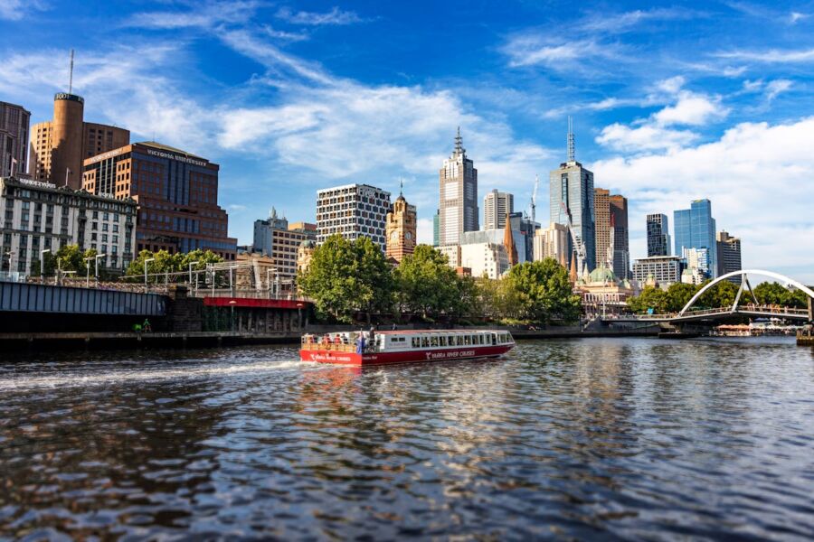 Melbourne river scene with skyline, skyscrapers, boat tours, greenery, bridges under sunny sky.