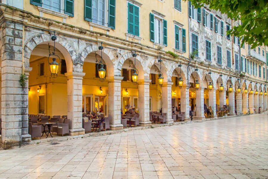 Mediterranean street with arched columns, green shutters, lanterns, outdoor seating, and stone tiles.