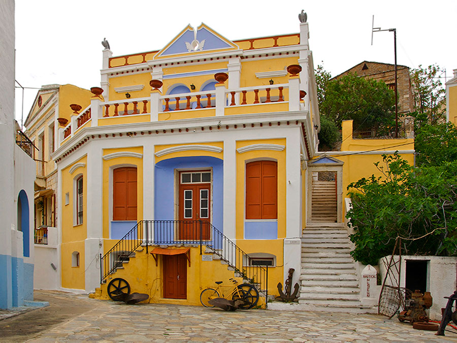 Yellow coastal naval museum facade with anchors, bicycle, neoclassical balcony in sunlit courtyard.