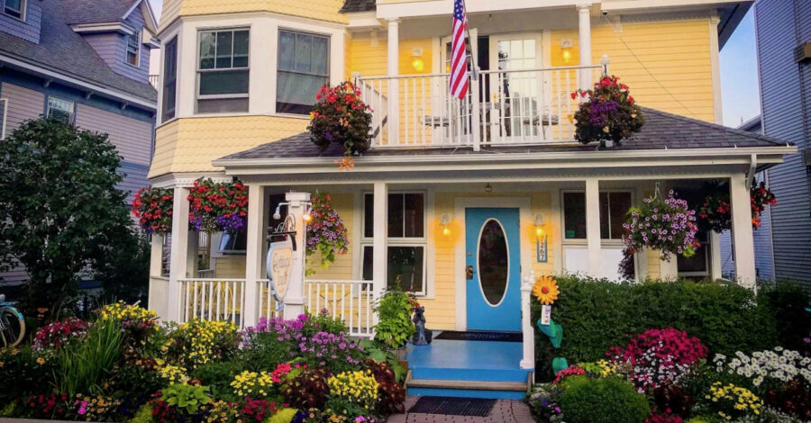 Charming yellow coastal inn with turquoise door, American flag, balcony, and colorful front garden.