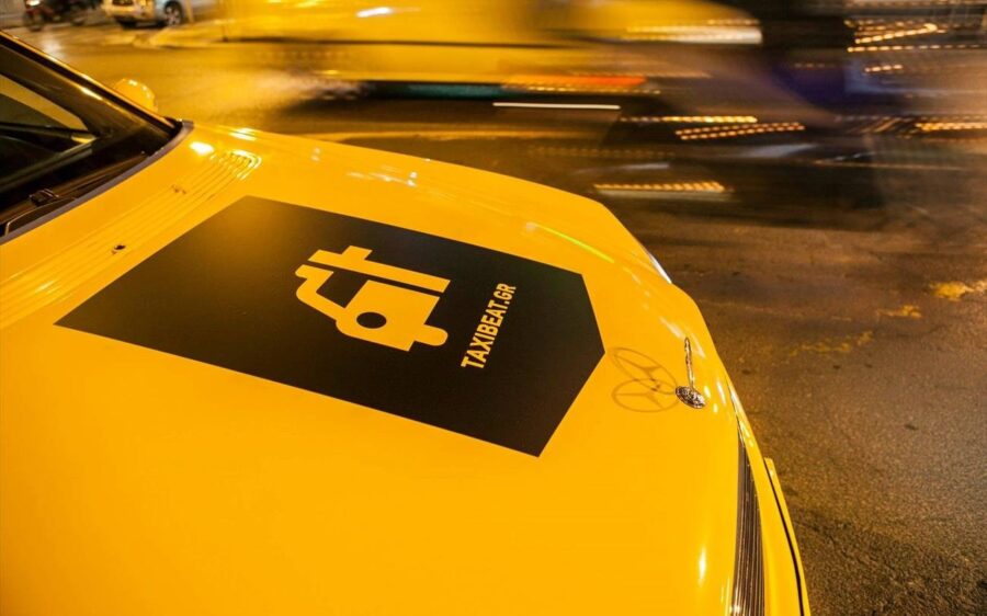 Yellow TAXIBEAT taxi roof in a bustling city at night with blurred lights.
