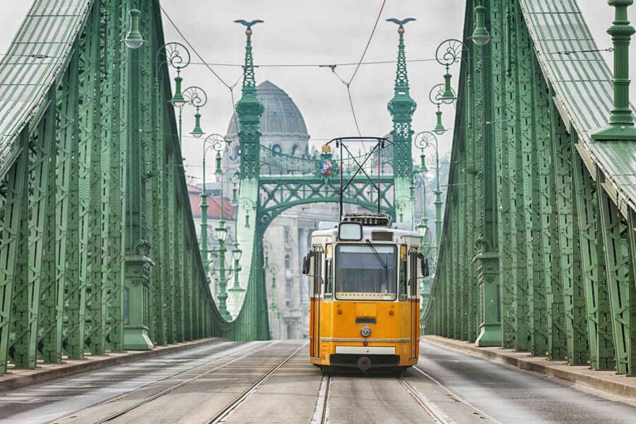 Yellow tram crossing ornate green bridge in Budapest with historical architecture in the background.