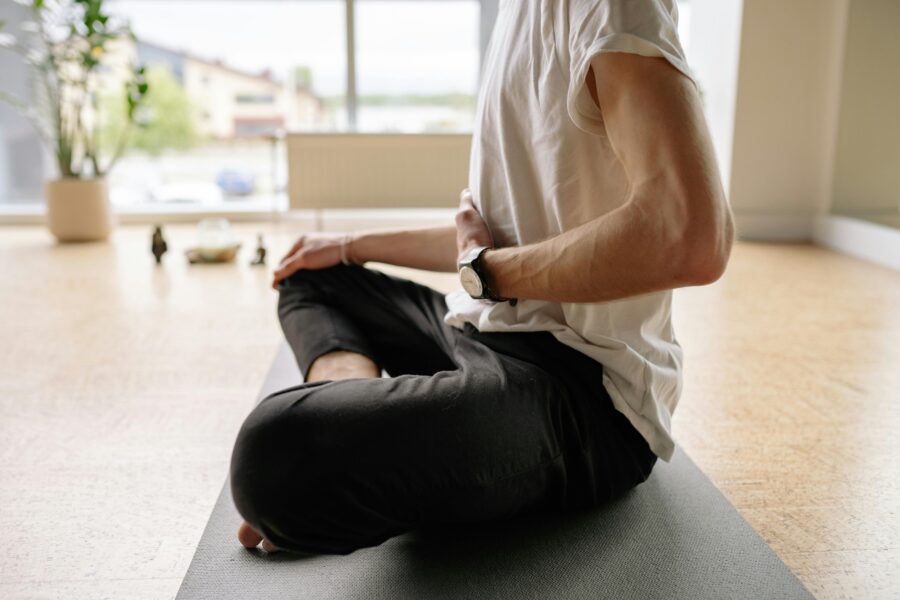 Man meditating indoors, cross-legged on yoga mat, surrounded by natural light and serenity.