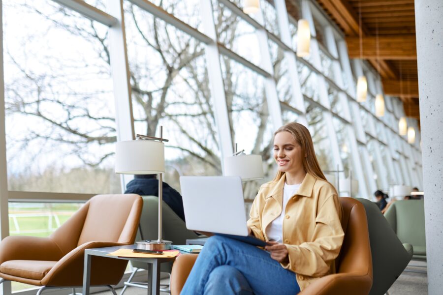 Happy young student woman studying on laptop in bright coworking lounge.