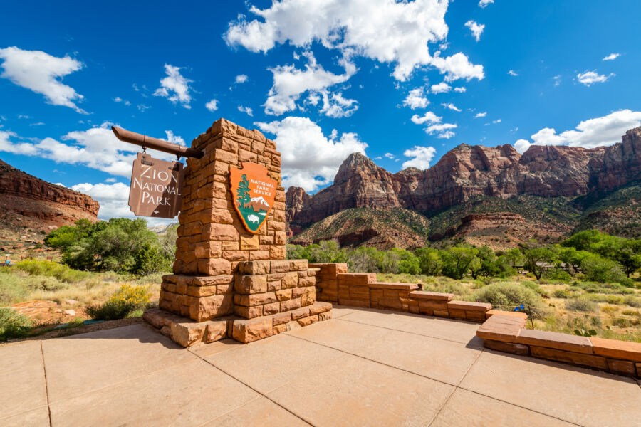 Entrance sign for Zion National Park displaying the park's name and emblem, surrounded by stunning red rock scenery