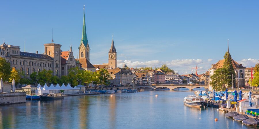 Picturesque Zurich waterfront with historical and contemporary architecture, blue sky, boats, and bridge.